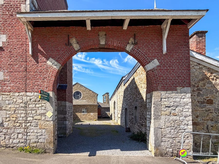 Backsteinbogen mit hölzernem Vordach, dahinter ein Hof mit Bruchsteingebäuden.