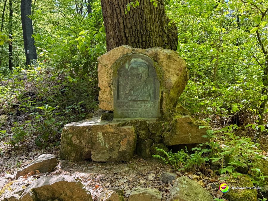 Steinernes Denkmal mit Relieftafel unter einem Baum im Wald.