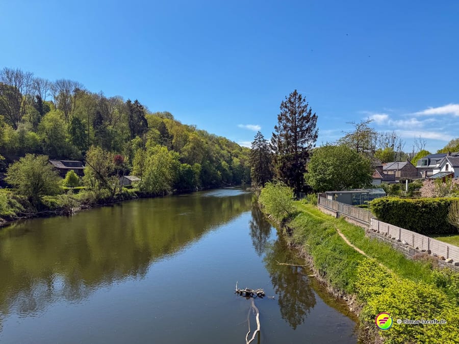 Der Fluss zwischen grasbewachsenen Ufern, dahinter bewaldete Hänge.