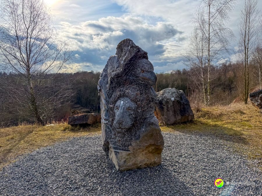 Moderne Steinskulptur oder Gedenkstein auf Kiesplatz vor bewölktem Himmel. Die verwitterte Formation könnte ein Denkmal für die Bergbaugeschichte der Region darstellen.