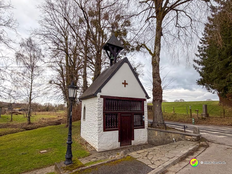 Kleine weiße Wegekapelle mit roter Holztür, Glockenturm und Kreuz. Die gepflegte Feldkapelle mit traditioneller Laterne dient als religiöser Rastplatz am Wanderweg.