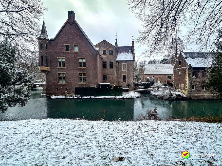 Laufen entlang eines verschneiten Ufers mit historischen Backsteinbauten und einem Wassergraben in einer ruhigen Winterlandschaft.