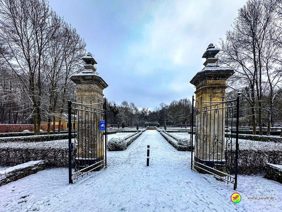 Laufen auf verschneitem Weg durch ein geöffnetes Tor mit steinernen Säulen in einem winterlichen Park mit kahlen Bäumen.