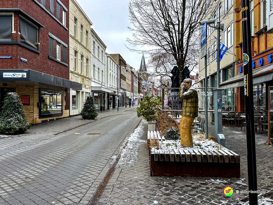 Laufen durch eine ruhige, gepflasterte Einkaufsstraße in Heerlen mit einer Holzskulptur auf einer Bank und winterlicher Atmosphäre.
