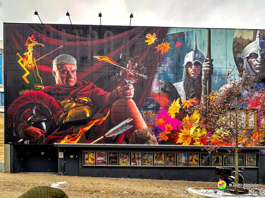 Laufen in Heerlen vorbei an einer großen, farbenfrohen Wandmalerei mit römischen Soldaten und herabfallenden Herbstblättern in einer urbanen Umgebung.