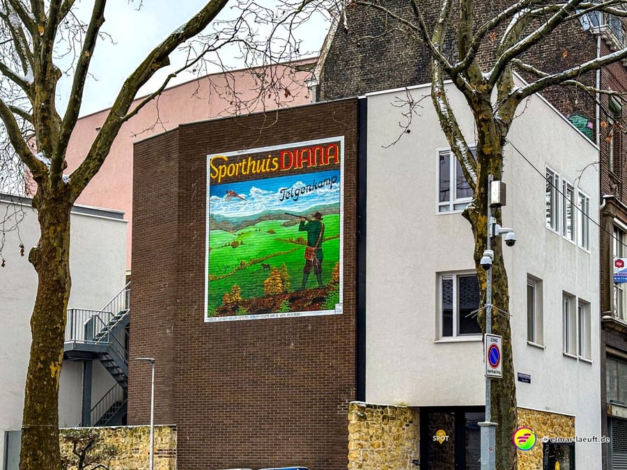 Laufen in Heerlen an einem urbanen Abschnitt mit einem Wandbild eines Jägers auf einem Gebäude zwischen kahlen Bäumen.