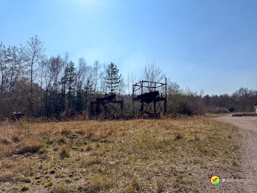 Laufen auf einem abgelegenen Weg in Maasmechelen mit verwildertem Gras und alten, rostigen Industrieanlagen am Waldrand unter klarem Himmel.