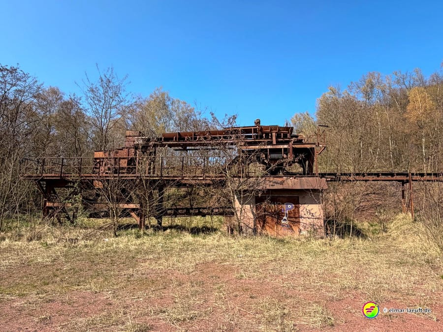 Laufen entlang einer verlassenen, rostigen Industrieanlage in einer offenen, von Bäumen umgebenen Landschaft bei klarem Himmel.