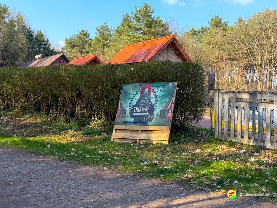 Laufen auf einem Waldweg in Maasmechelen mit einem auffälligen Schild eines Clowns, das den Weg weist, vor einer Hecke und kleinen Häusern mit roten Dächern.